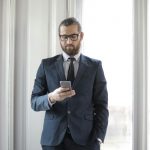Confident man in formal suit using smartphone by window in office setting.