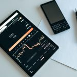 Top view of a workspace with a tablet displaying financial charts, a smartphone, pen, and paper.