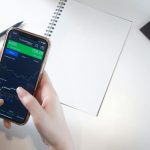 Close-up of hands using smartphone stock market app with notebook and pencil on desk.