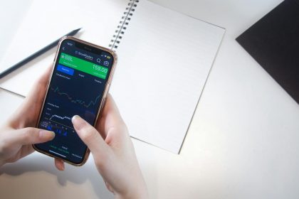Close-up of hands using smartphone stock market app with notebook and pencil on desk.